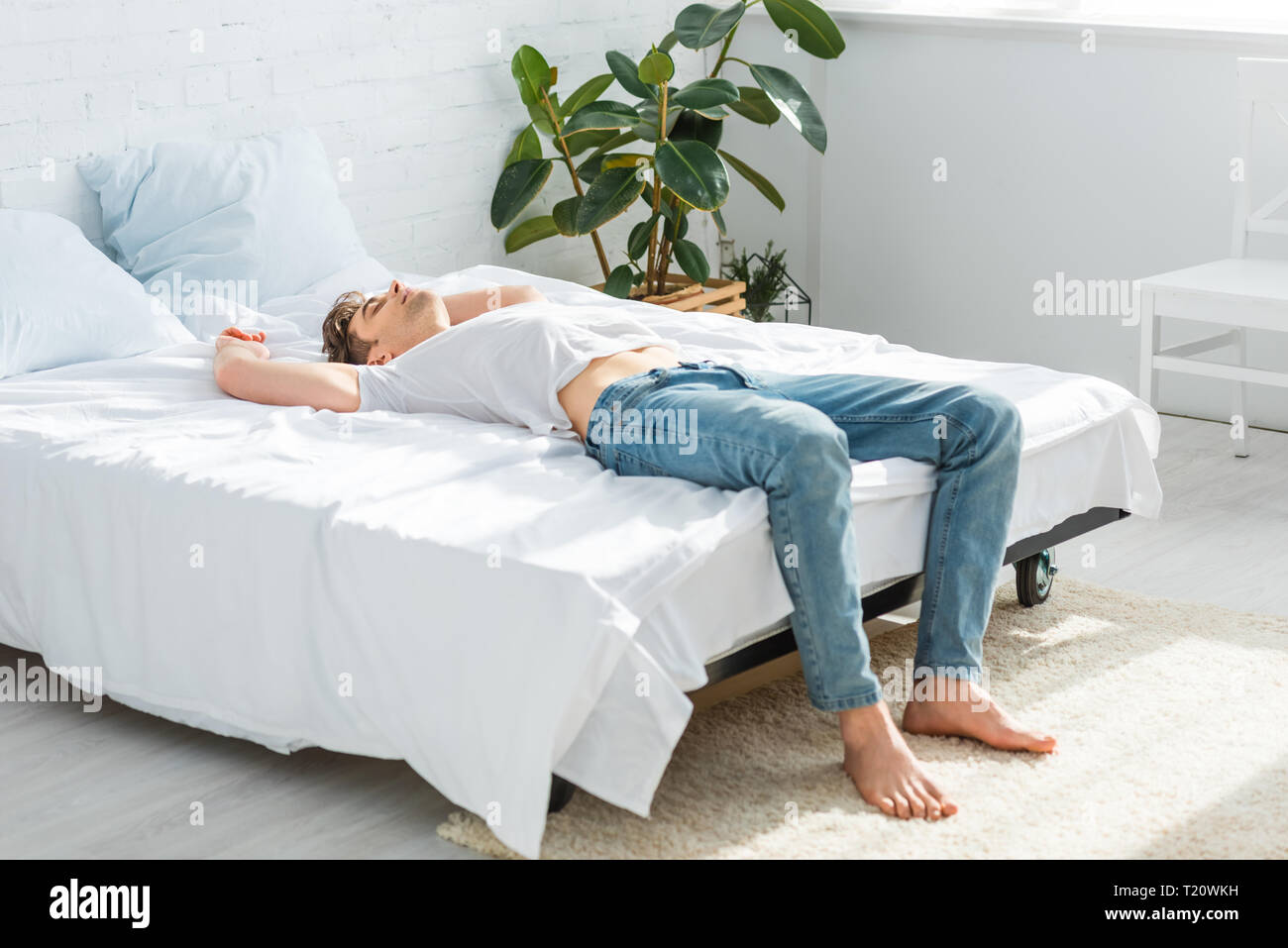 Uomo bello in maglietta e jeans giacente sul retro sul letto in camera da letto Foto Stock