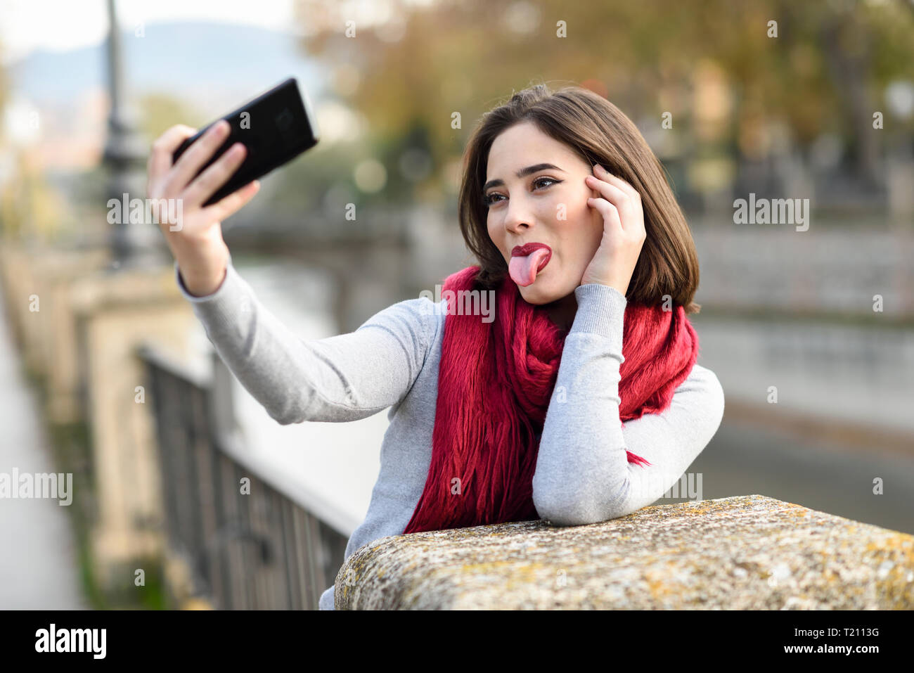 Ritratto di giovane donna che indossa sciarpa rossa spuntavano lingua tenendo selfie con un telefono cellulare Foto Stock