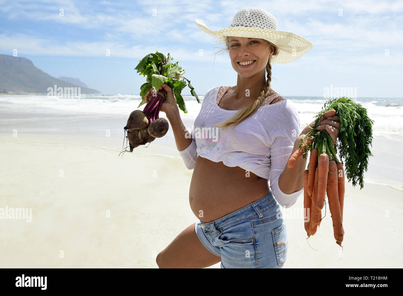 Ritratto di sorridere donna incinta sulla spiaggia tenendo le carote e barbabietole Foto Stock