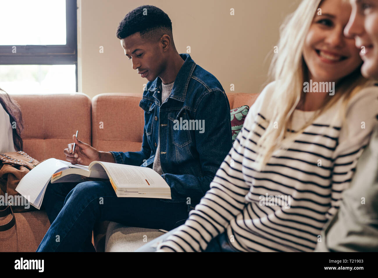 Maschio africano studente rendendo note durante la lettura di libri seduti al divano presso il college campus. Studente presso il campus edificio con le ragazze in seduta fro Foto Stock