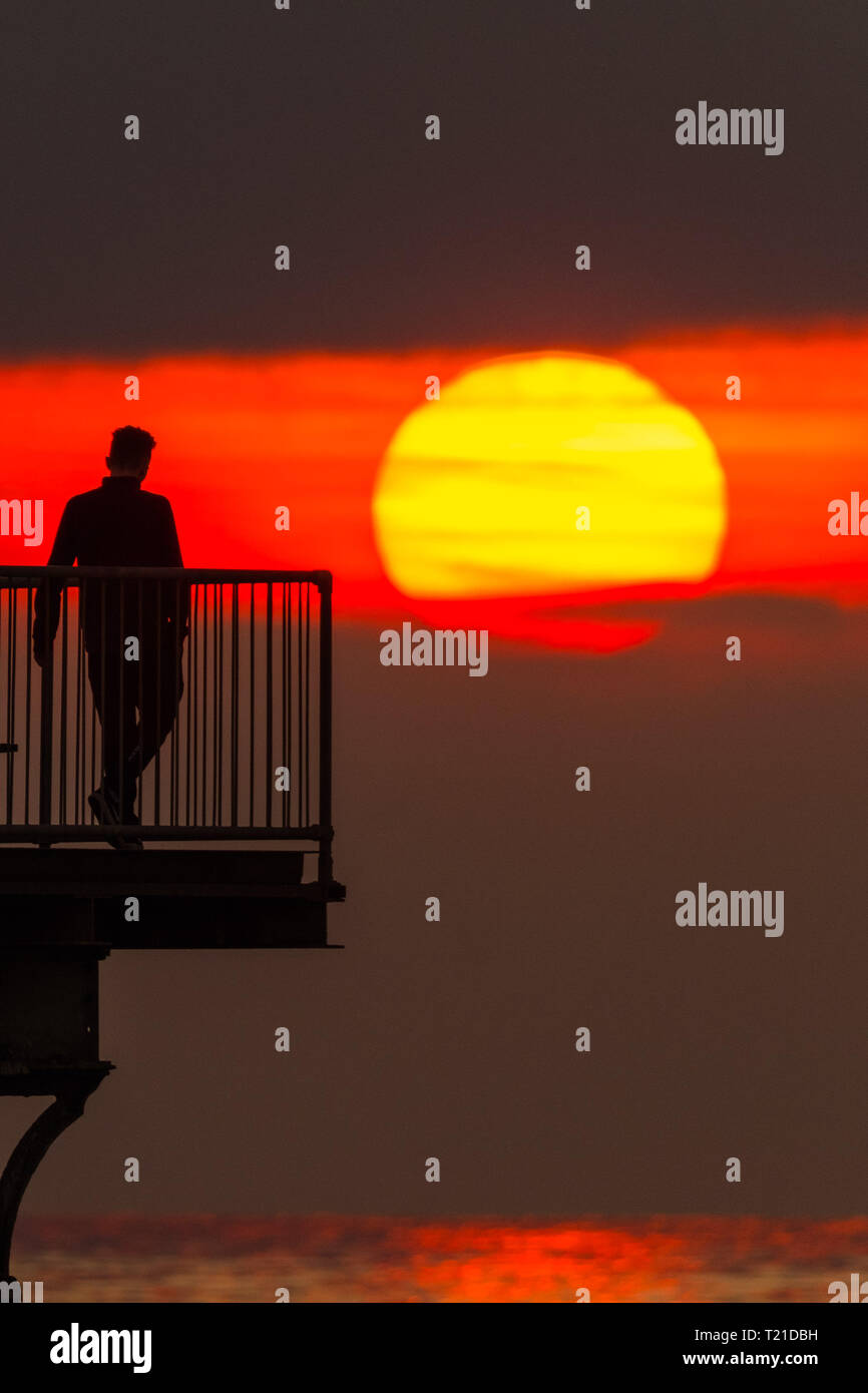 Aberystwyth, Galles. 29 mar 2019. Regno Unito Meteo: un uomo gustando un drink serale alla fine del molo a Aberystwyth, il Cardigan Bay costa del Galles occidentale si stagliano dall'impostazione fiammeggiante sun . Alta pressione continua a dominare il meteo per molto di Inghilterra e Galles, con si stabilirono le condizioni meteo per durare almeno un altro giorno , prima di più condizioni dello scambiatore di calore di ritorno. Foto:Keith Morris/Alamy Live News Foto Stock