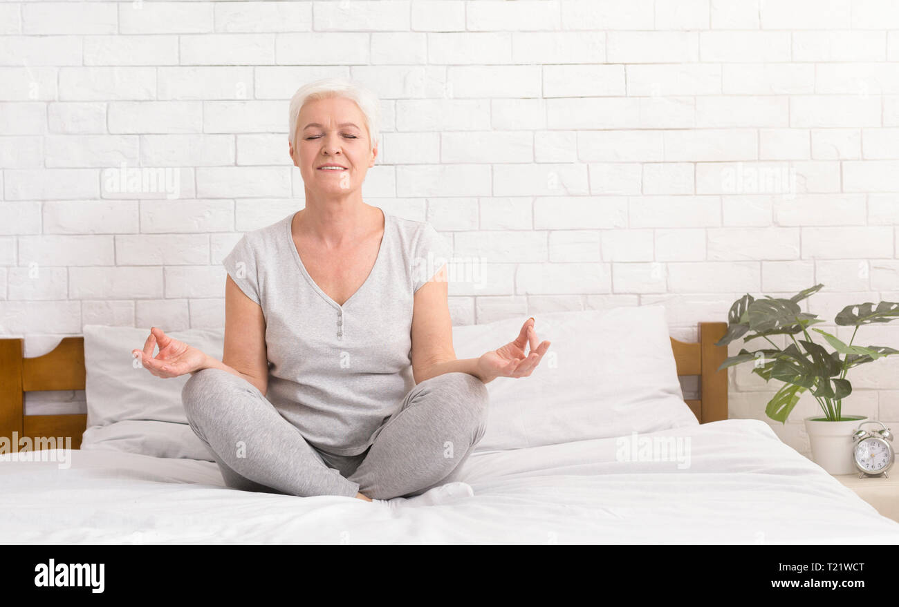 La meditazione. Senior donna meditando nel letto, seduti nella posizione del loto e la pratica di anti-stress lo yoga, spazio libero Foto Stock