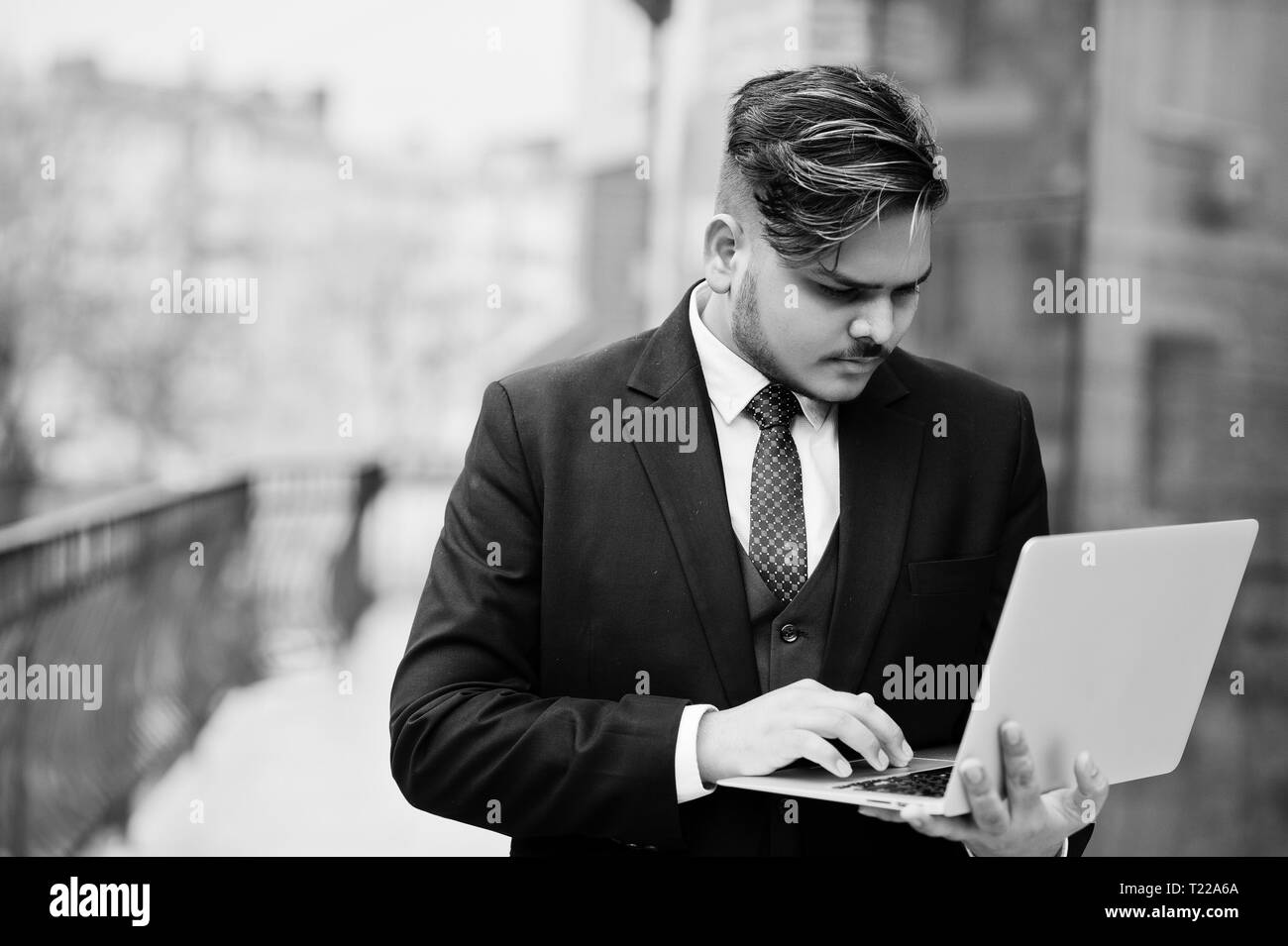 Elegante imprenditore indiano in abbigliamento formale con il computer portatile su mani in piedi contro windows nel centro business. Foto Stock