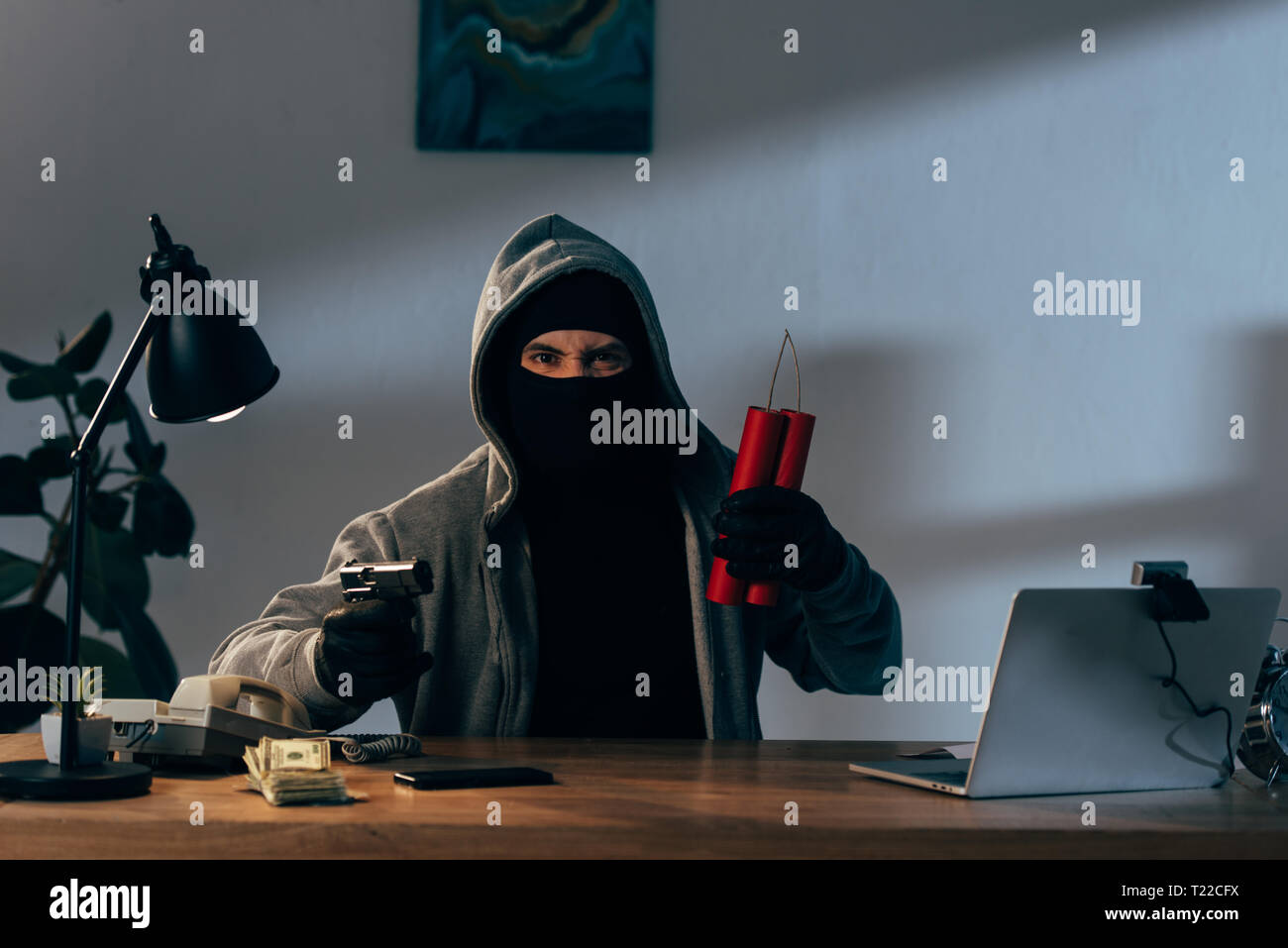 Arrabbiato terrorista in maschera tenendo la pistola e la dinamite mentre è seduto alla scrivania in camera oscura Foto Stock