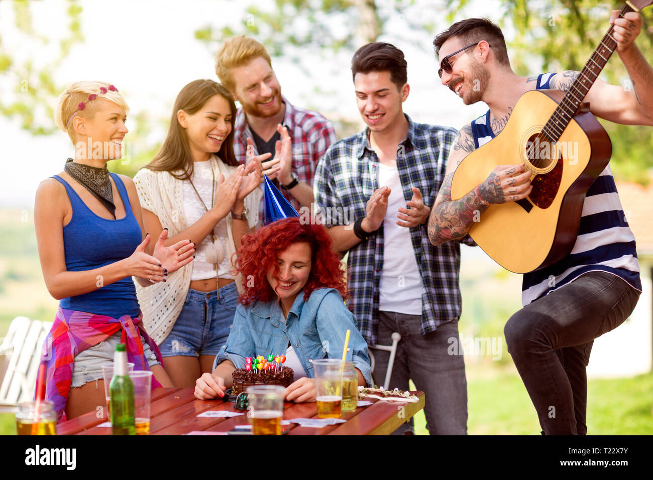 Timido curly redhead compleanno ragazza con gli amici che applaudire e riproduci canzone di buon compleanno alla chitarra per esterno Foto Stock