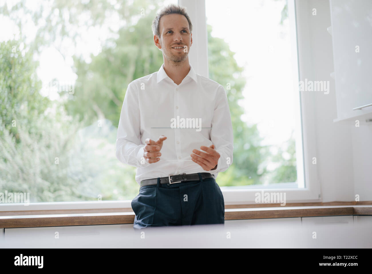 Imprenditore in piedi alla finestra della cucina, utilizzando vetro touch screen, sorridente Foto Stock