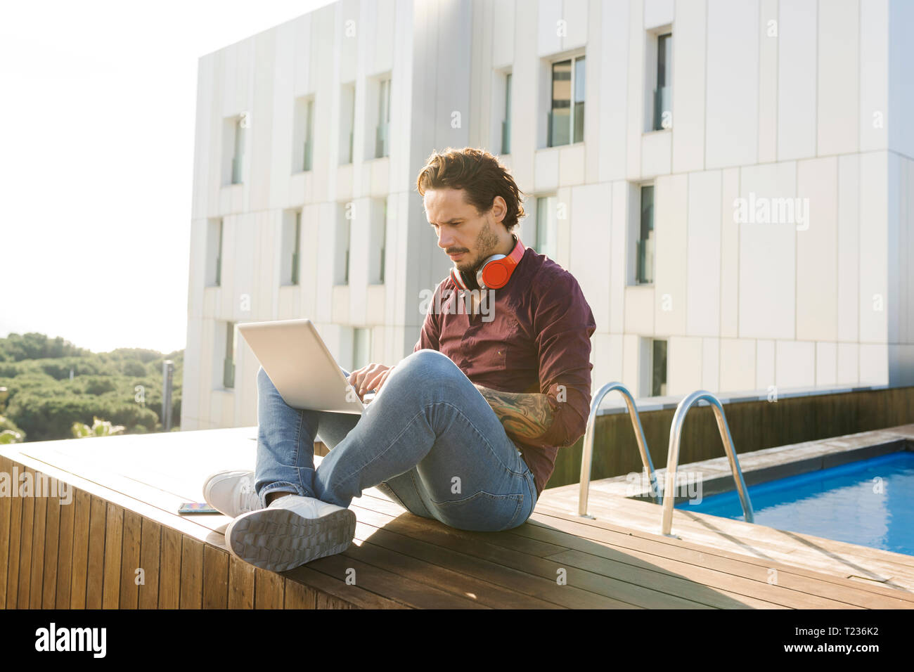 Uomo seduto a gambe incrociate su una terrazza sul tetto, utilizzando laptop, indossando le cuffie Foto Stock