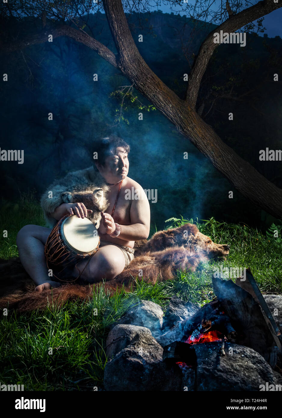 Caveman vestito in pelle di lupo suonare il tamburo vicino al falò in foresta Foto Stock