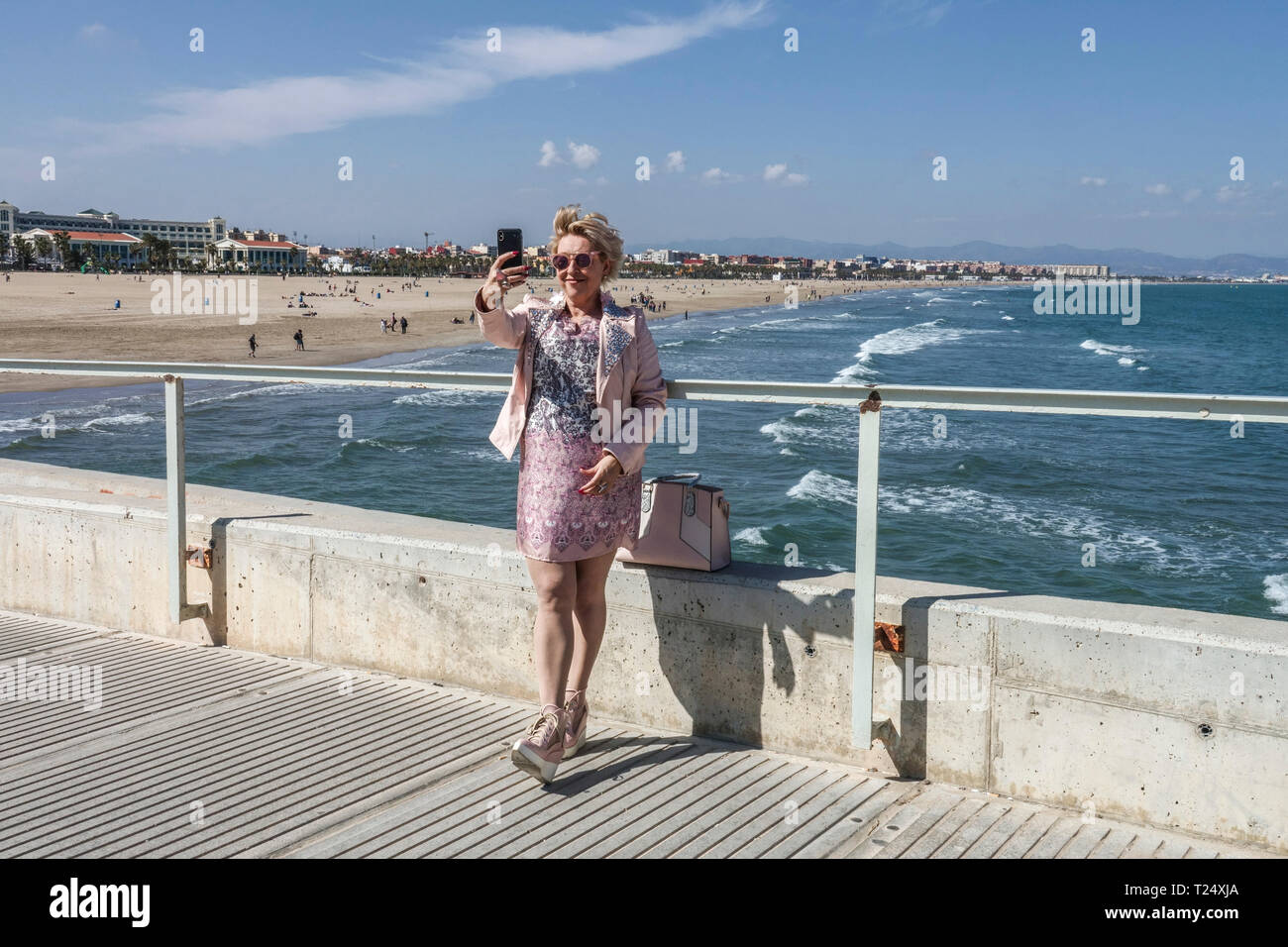 Una donna in un abito beige prendendo un selfie su un molo di cemento con mare e spiaggia Malvarrosa Valencia vista mare Valencia Spagna spiaggia Foto Stock