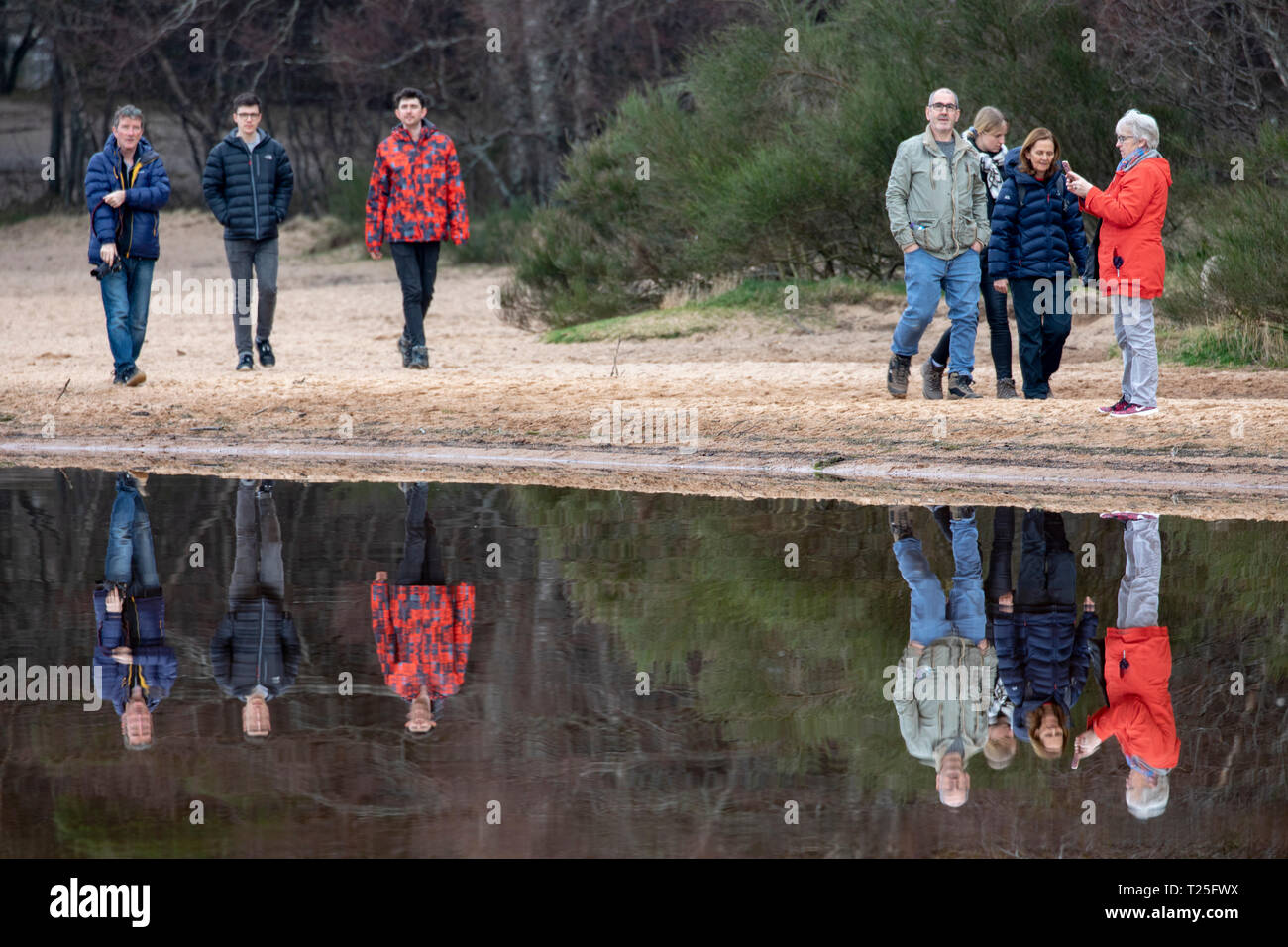 Le persone che si godono una passeggiata lungo il litorale si riflettono nelle tranquille acque del Loch Morlich vicino Aviemore nelle Highlands scozzesi Foto Stock