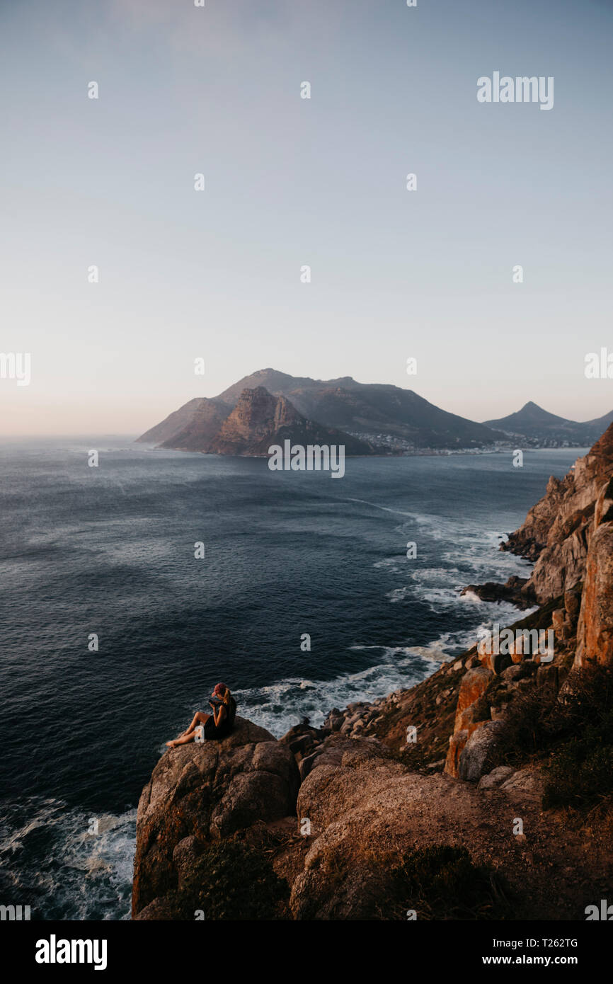 Sud Africa, Western Cape, donna seduta su una roccia tramite telefono cellulare, visto da Chapman's Peak Drive Foto Stock