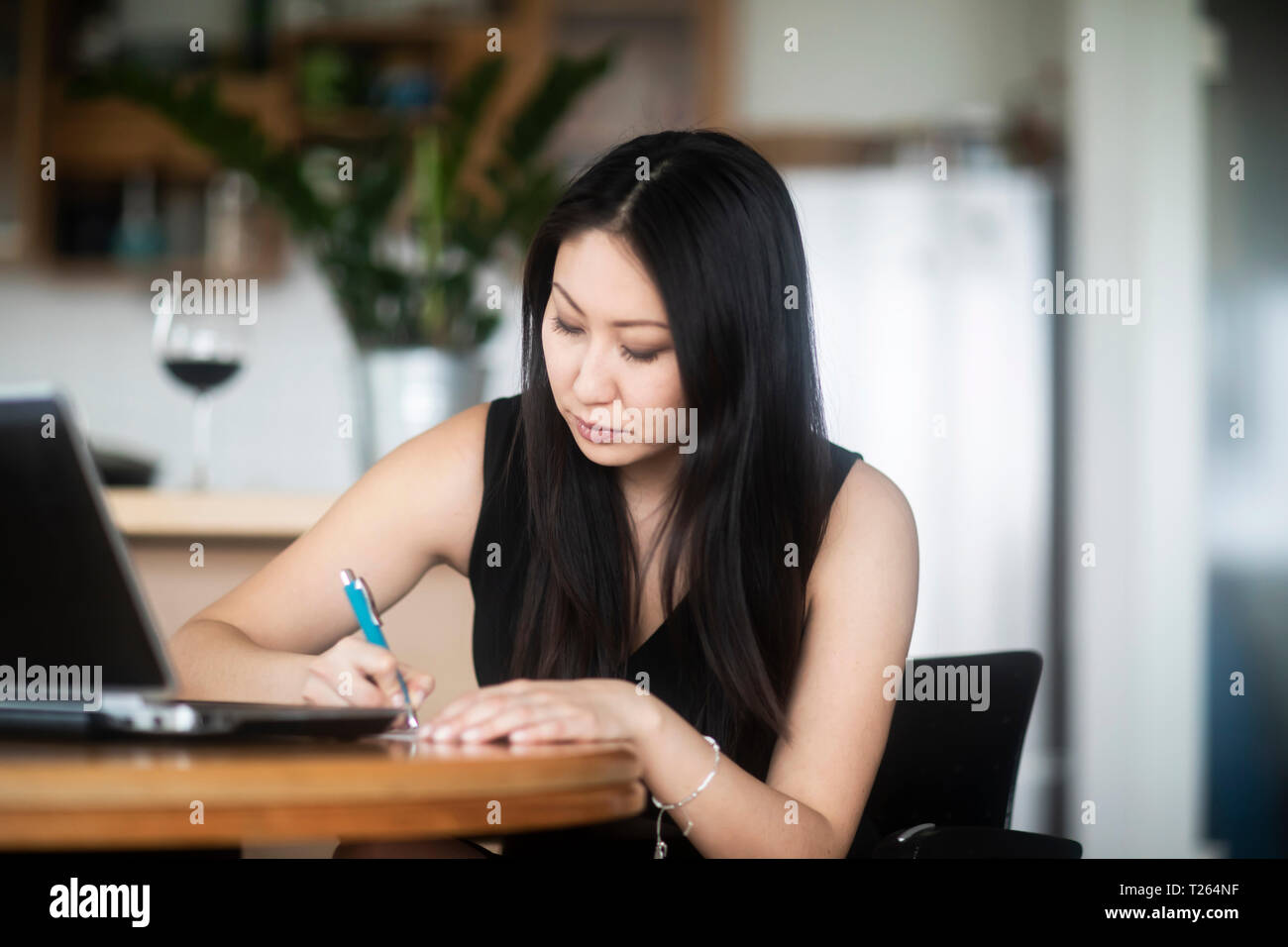 Giovane donna in abito nero utilizzando laptop e prendere appunti sul tavolo a casa Foto Stock