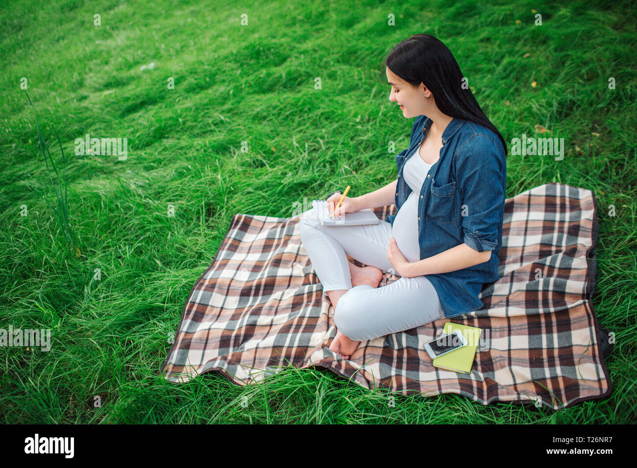 Ritratto di un felice capelli neri e orgogliosa donna incinta nel parco. Il modello femminile è seduto sull'erba ed è scrivere note Foto Stock