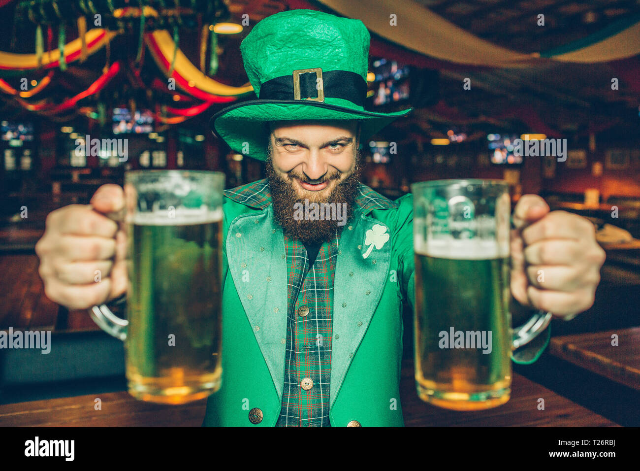 Felice e eacited giovane san Patrizio tuta in pub da solo. Egli tenere due boccali di birra e guardare su telecamera Foto Stock