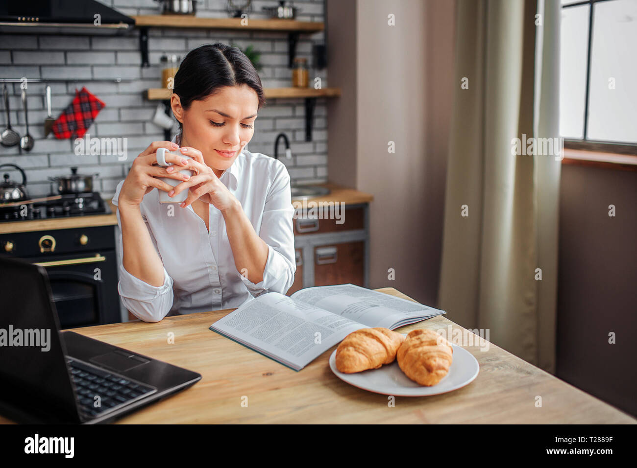 Intelligente donna adulto seduto a tavola in cucina. Ha letto ufficiale e tenere premuto tazza bianca con bevanda calda. La donna guarda verso il basso. Ella lavora a casa Foto Stock