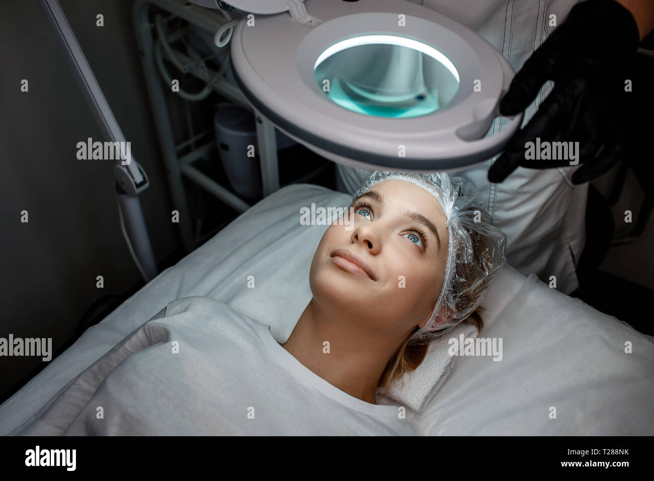 Giovane donna nel cappuccio sdraiato sul lettino in camera. Lei guarda alla lampada al di sopra di lei. Modello sorriso. Beautisian stand al suo capo e spostare la lampada con le mani. Foto Stock