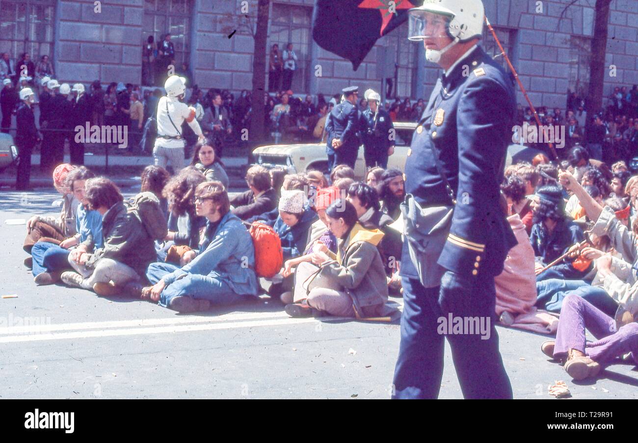 Durante il 1971 giorno di maggio le proteste contro la guerra in Vietnam, un gruppo di vestiti caldamente maschio e femmina di contestatori siedono insieme nel mezzo del decimo St NW, in una giornata di sole, un off-telecamera onde figura un nero, rosso e verde 'parte internazionale della gioventù' o 'Yippie' bandiera a Washington DC, maggio 1971. () Foto Stock