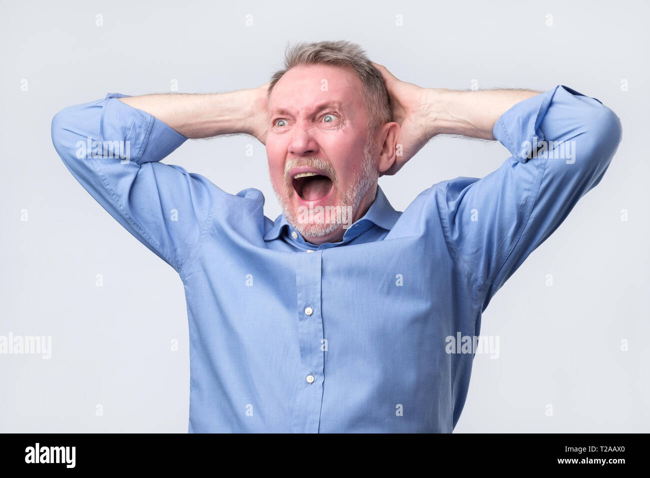 Senior Uomo in camicia blu con arrabbiato grimace sul suo viso,con la bocca aperta a gridare, pronto a discutere. Studio shoot. Negativo emozione umana. Foto Stock