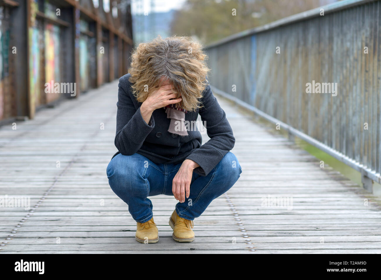 Sconsolato donna accovacciata su un ponte appeso la sua testa con la mano al suo fronte all'aperto in un freddo giorno Foto Stock