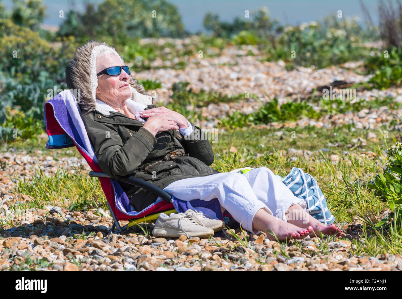 Senior donna che indossa un cappotto seduto su una sdraio sulla spiaggia in un freddo giorno di primavera nel Regno Unito. Foto Stock