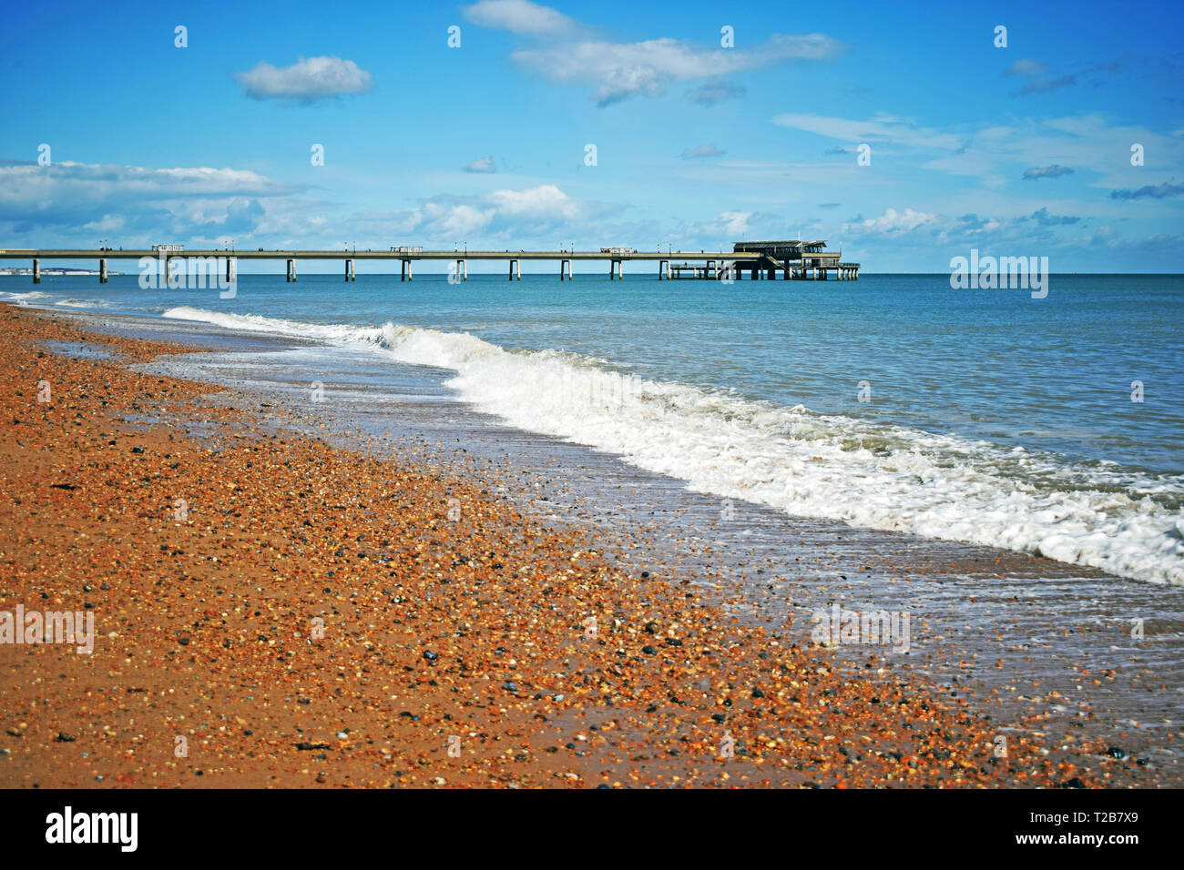 Mare di scena a trattare in Kent England con le onde provenienti sulla spiaggia di ciottoli e il molo che si allunga nel mare blu acqua con un cielo nuvoloso sopra Foto Stock