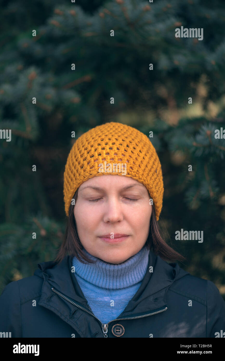 Bella donna di 40s headshot ritratto nel parco con gli occhi chiusi, rilassato e informale persona femmina con cappuccio giallo in posa davanti ad abete di sunny s Foto Stock