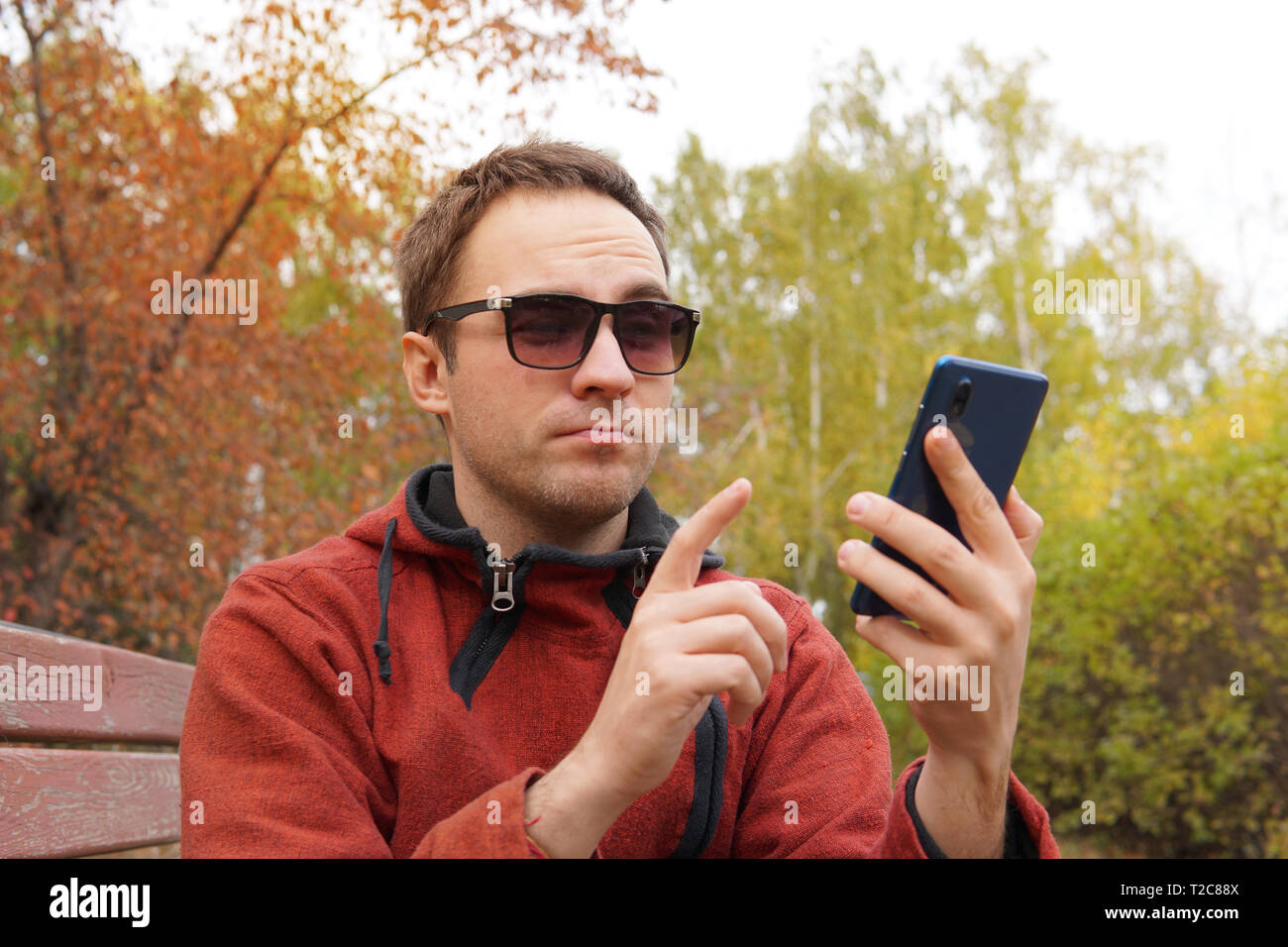 Guy europea si siede su una panchina di una città parco d'autunno. Un uomo è sorridente guardando il telefono. concetto di gioia e piacere, gioia e allegria. Umorismo, organi giurisdi Foto Stock