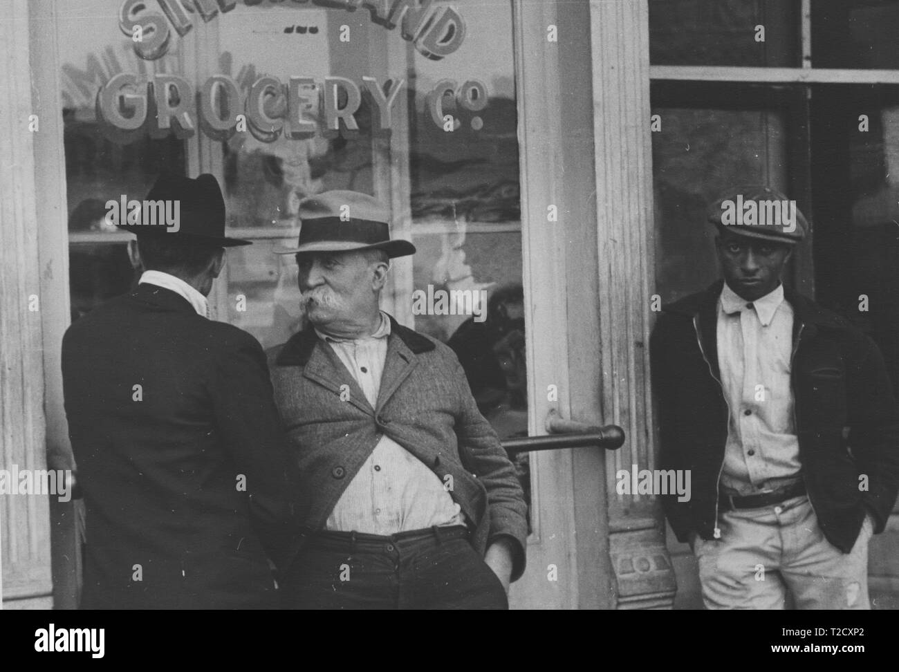 Fotografia in bianco e nero di tre uomini stavano in piedi davanti al 'Smithland drogheria Co, ' tra cui un uomo maturo con un grandi baffi bianchi e una maglietta sporca, che chat con un uomo che indossa un cappello e una tuta, mentre un terzo uomo, indossando una leggermente inclinata cap e un buio, zipper jacket, si appoggia contro un pilastro a destra, guardando verso la fotocamera con un espressione seria sul suo volto; situato in Smithland, Kentucky, Stati Uniti d'America; fotografata da Ben Shahn, sotto il patrocinio degli Stati Uniti " Farm Security Administration in 1935. Dalla Biblioteca Pubblica di New York. () Foto Stock