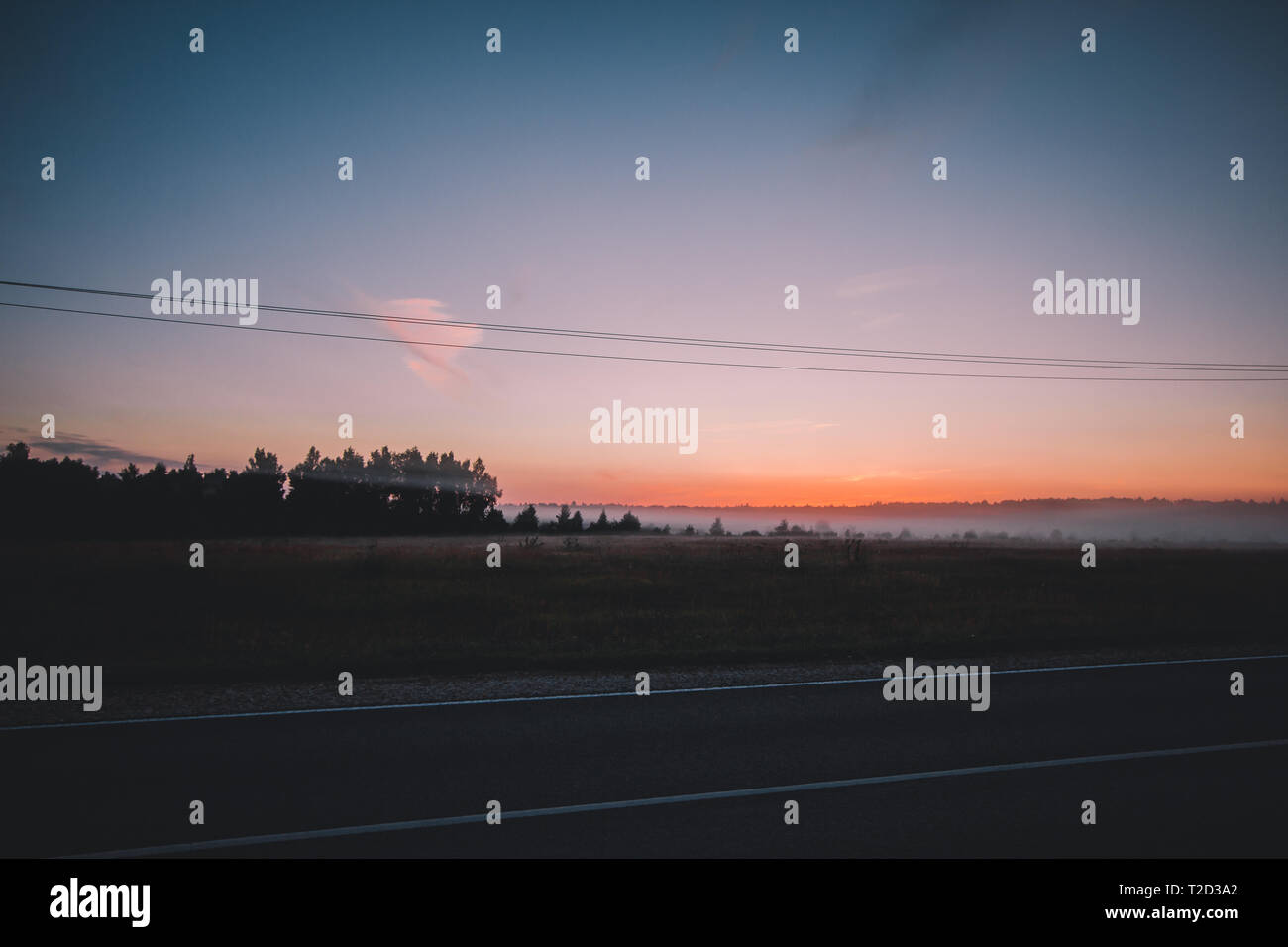 Mattina è venuto. Il sole arancione aumenta al di sopra di un campo di misty, strada asfaltata e la foresta di alberi scuri. Una fitta nebbia si chiude la strada alla luce del sole per i foggy gr Foto Stock