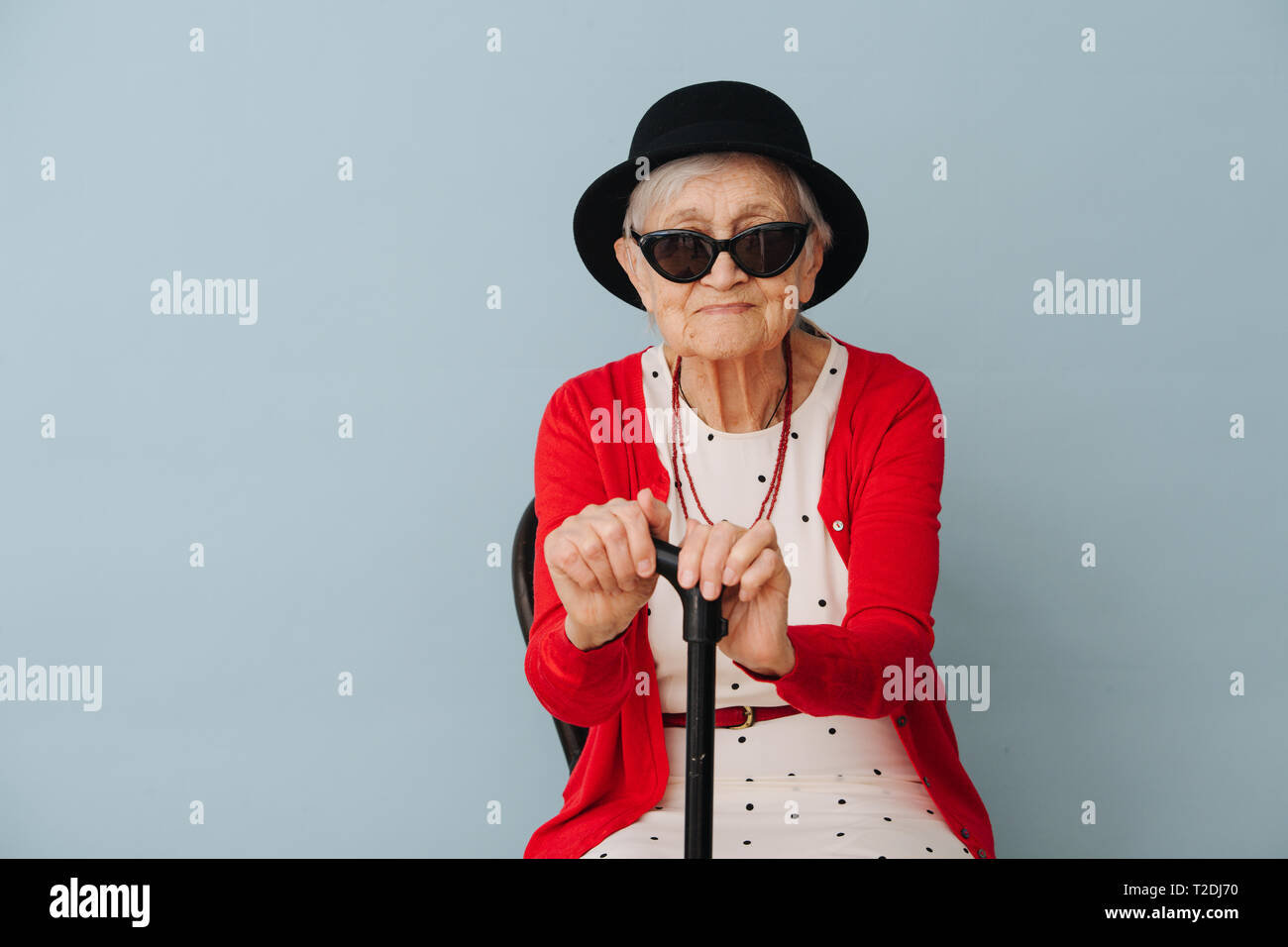 Rispettabile ben vestita di nero mini cappello e occhiali da sole dai capelli grigi di fantasia donna senior con la canna da zucchero è in appoggio su una sedia a casa su una soleggiata giornata di primavera Foto Stock