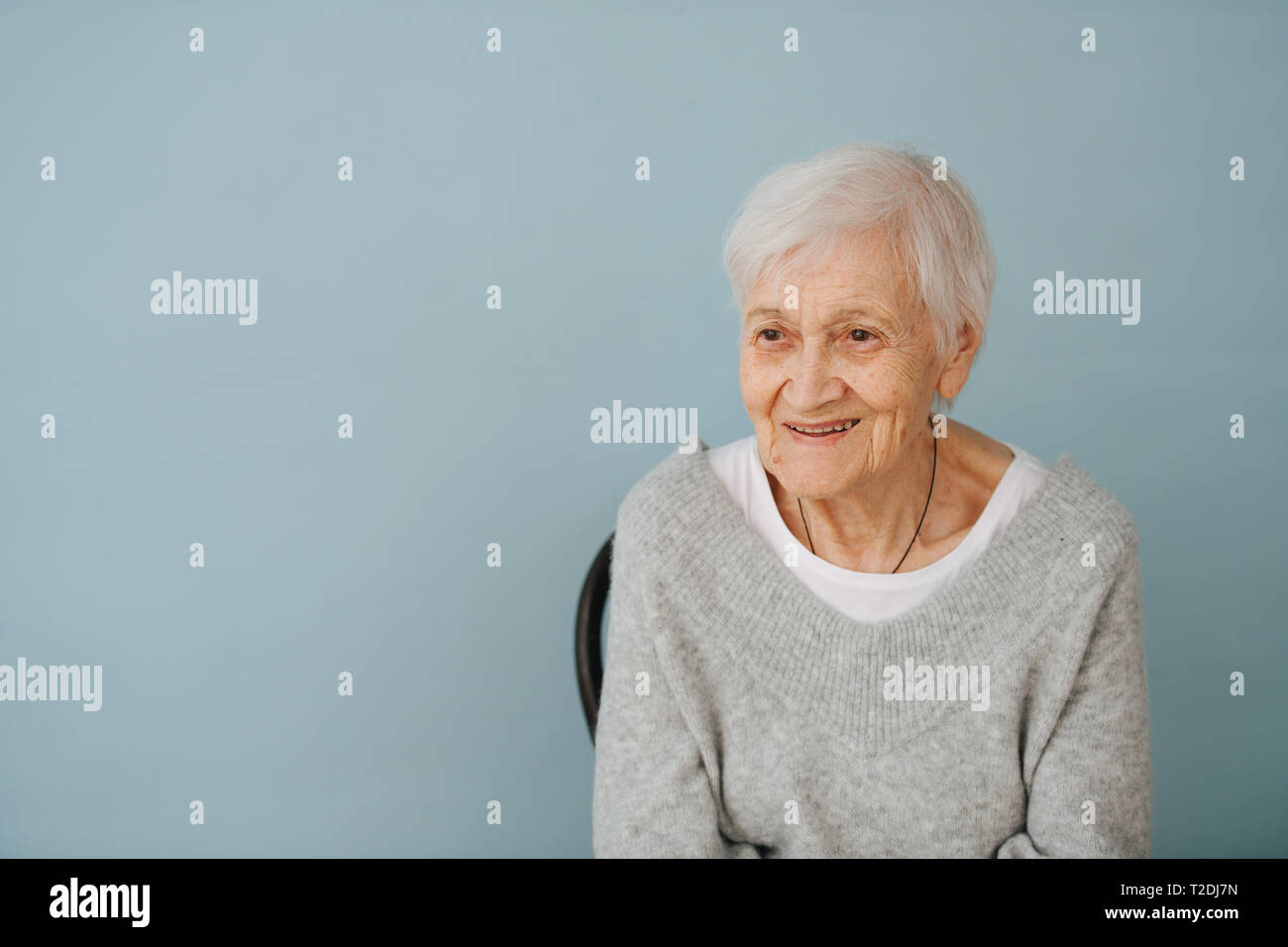 Ritratto di sorridere anziani dai capelli grigi donna seduta su una sedia, indossare comode grigio V-tipo maglione a casa. Occhiali off. Foto Stock