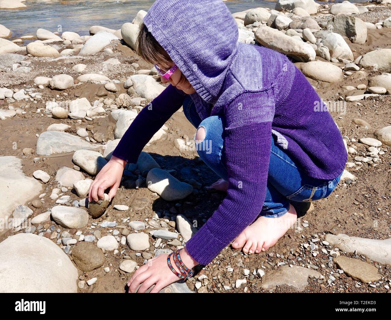 Carino dieci anno vecchia ragazza indossando felpa con cappuccio di colore viola e rosa occhiali, prelievo di rocce dal Fiume Ventura, in un ambiente rurale, Ojai, CALIFORNIA, STATI UNITI D'AMERICA Foto Stock
