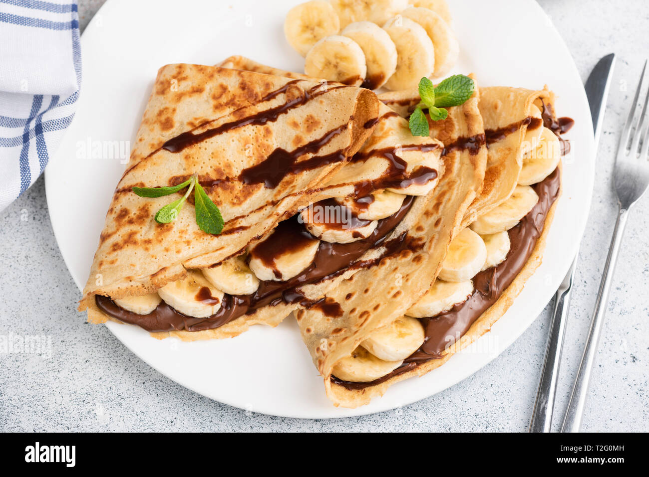 Crepes o blini farciti con cioccolato noccioletta, banana su una piastra bianca. Primo piano Foto Stock