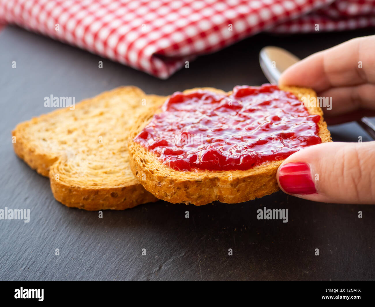 Una donna con le unghie dipinte di rosso con una fetta di pane tostato in mano con confettura di lamponi Foto Stock