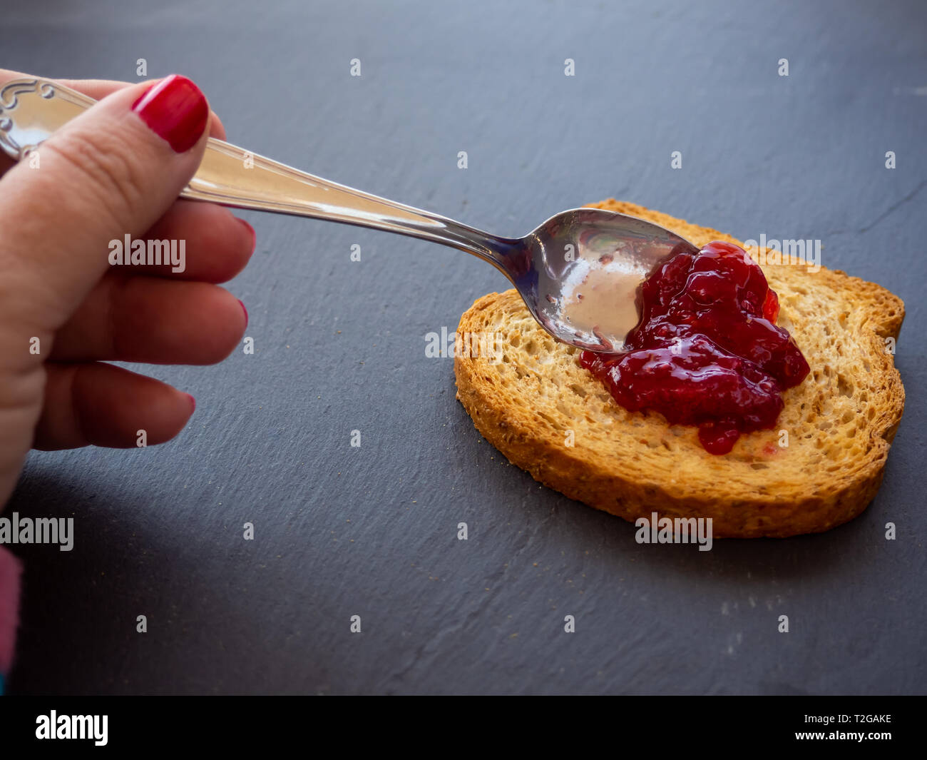 Una donna con unghie dipinte di rosso di spalmatura di marmellata di lamponi con un cucchiaio su una fetta di pane tostato Foto Stock