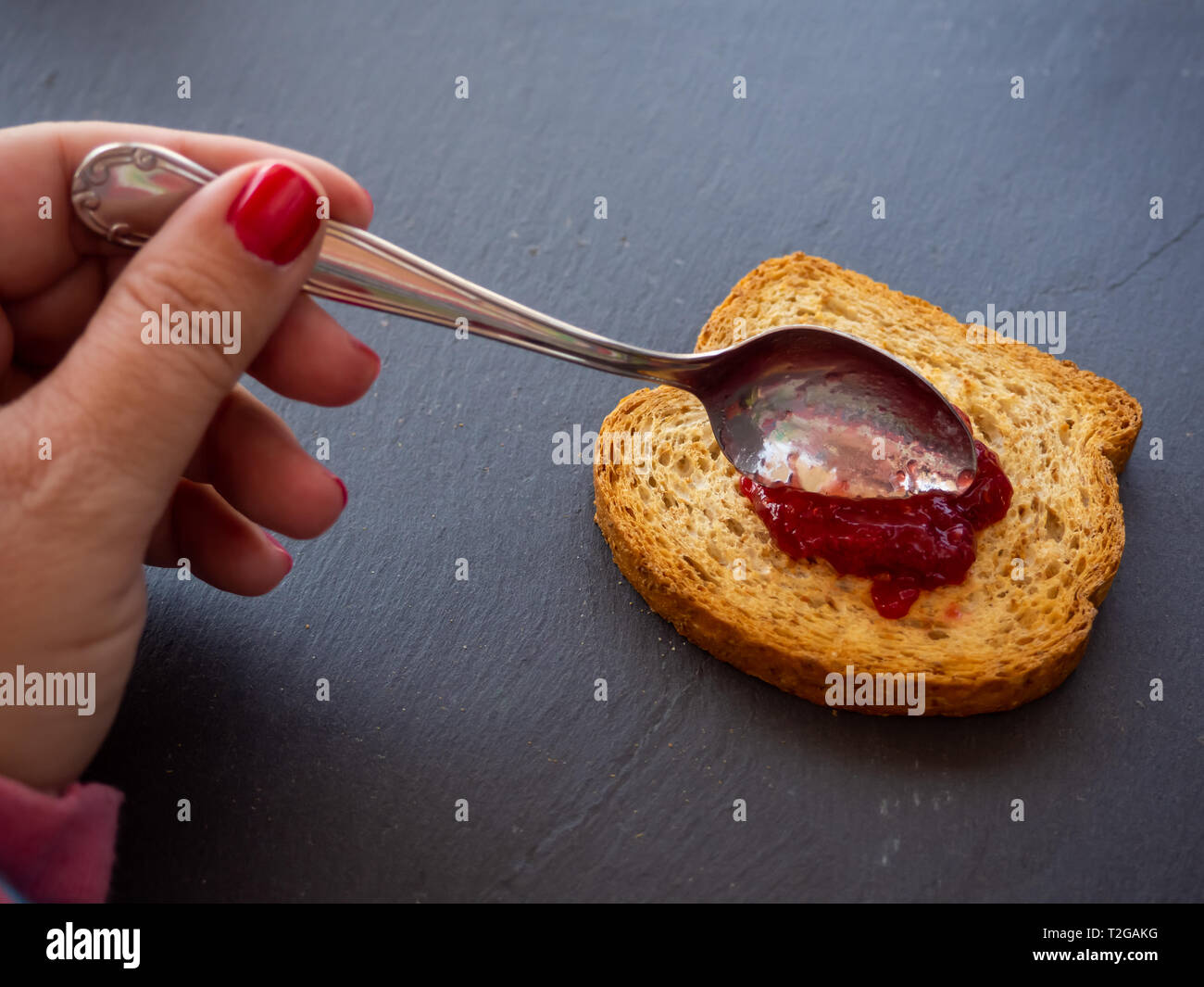 Una donna con unghie dipinte di rosso di spalmatura di marmellata di lamponi con un cucchiaio su una fetta di pane tostato Foto Stock