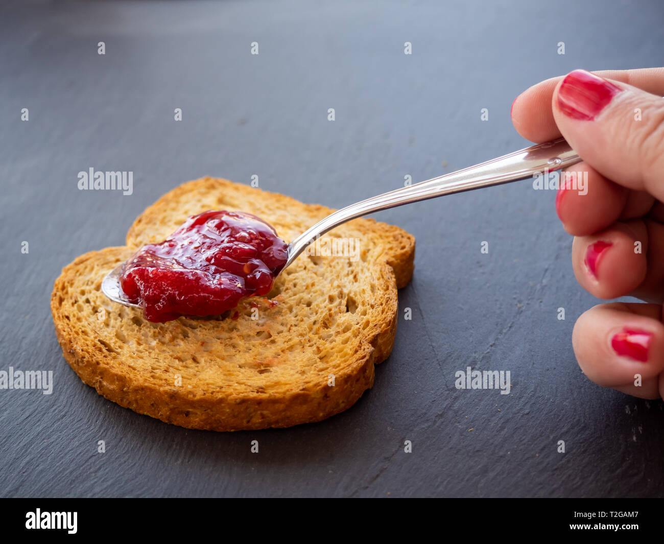 Una donna con unghie dipinte di rosso di spalmatura di marmellata di lamponi con un cucchiaio su una fetta di pane tostato Foto Stock
