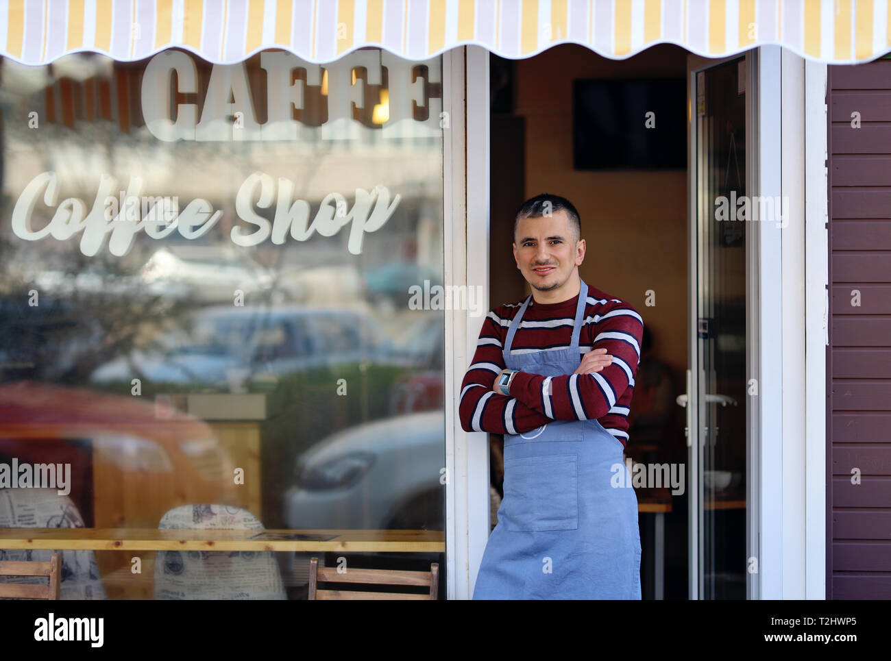 Un bel giovane coffee shop proprietario in piedi all'ingresso del suo negozio. Coffee shop iscrizione sulla finestra del negozio. Egli è un fiero proprietario del negozio Foto Stock