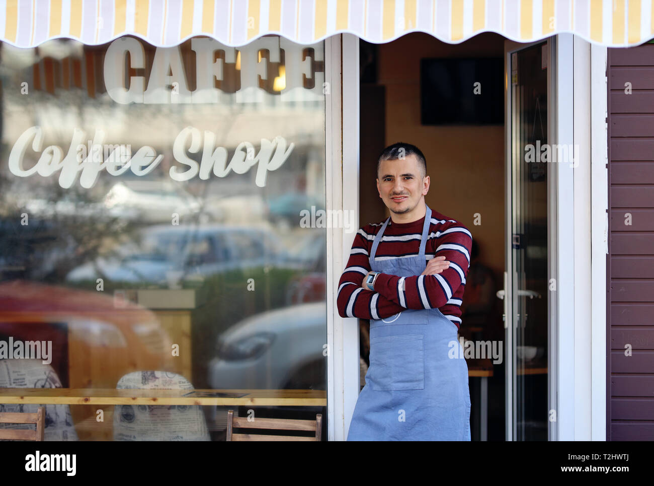 Un bel giovane coffee shop proprietario in piedi all'ingresso del suo negozio. Coffee shop iscrizione sulla finestra del negozio. Egli è un fiero proprietario del negozio Foto Stock