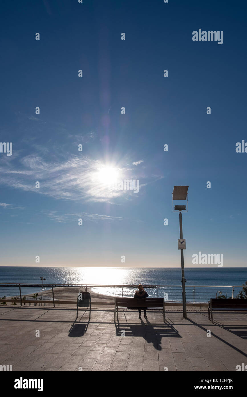 La donna si siede su un banco di lavoro godendo il forte sole invernale sul Mediterraneo presso il Mirador de la Plaza de Topete, città di Alicante, Spagna Foto Stock
