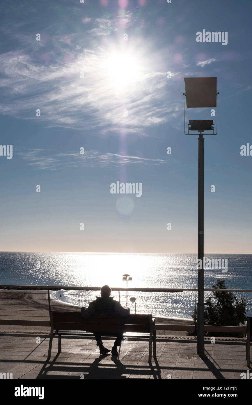 Uomo si siede su un banco di lavoro godendo il forte sole invernale sul Mediterraneo presso il Mirador de la Plaza de Topete, città di Alicante, Spagna Foto Stock