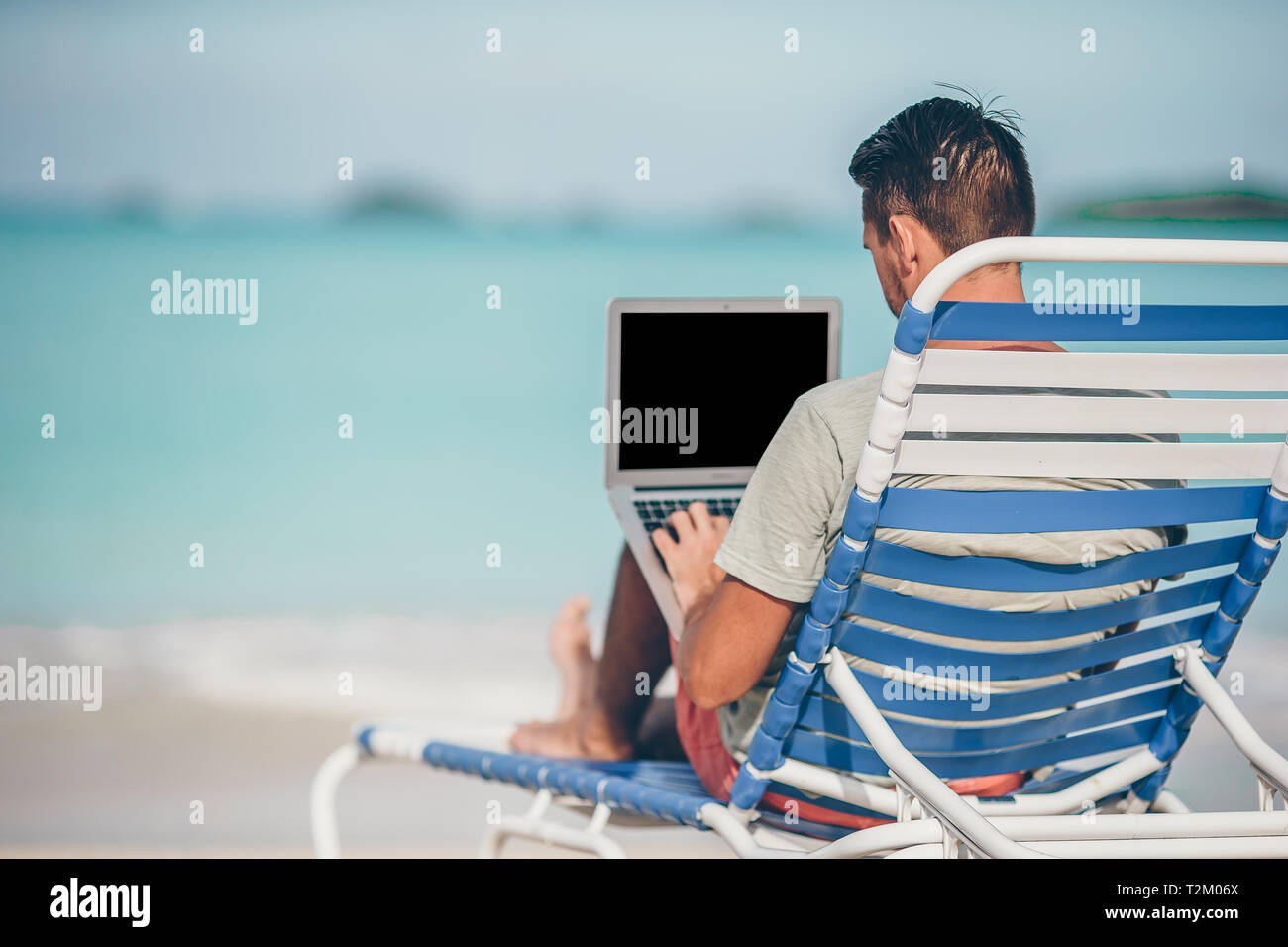 Giovane uomo seduto sulla sabbia con il computer portatile sulla tropicale spiaggia dei Caraibi. Uomo con computer e lavorare sulla spiaggia Foto Stock