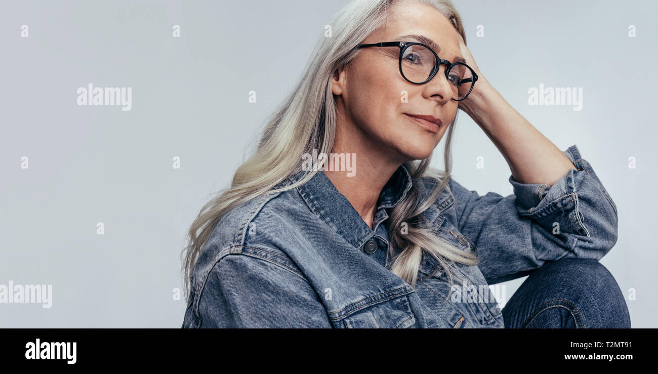 Senior caucasian donna seduta con la mano sulla testa e guardando lontano pensando. Femmina matura con occhiali contemplando contro uno sfondo grigio. Foto Stock