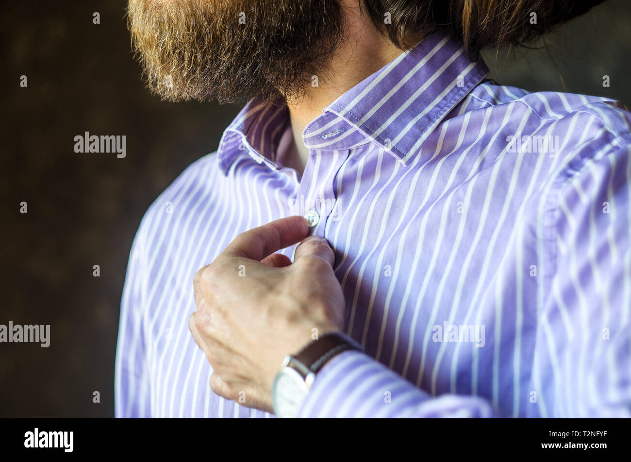 Primo piano del giovane uomo barbuto in orologio da polso abbottonatura suo Striped Shirt ottenere pronto ad andare fuori. Usura al concetto di lavoro. Foto Stock