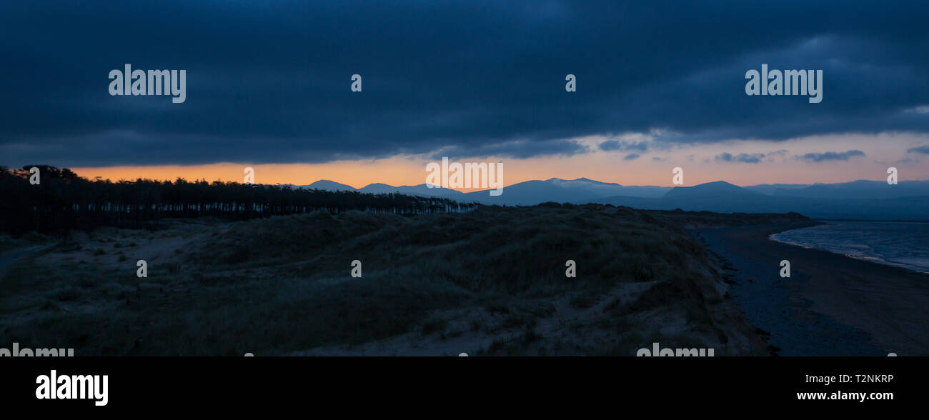 Vista panoramica di sunrise a Newborough Warren, Anglesey. Il sorgere del sole su Snowdonia montagne in distanza come dawn si rompe; nuvole scure, scuro litorale. Foto Stock