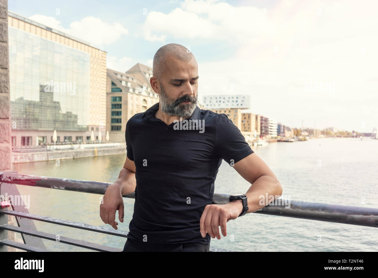 Uomo che guarda smartwatch sul ponte, sul fiume e gli edifici in background, Berlino, Germania Foto Stock