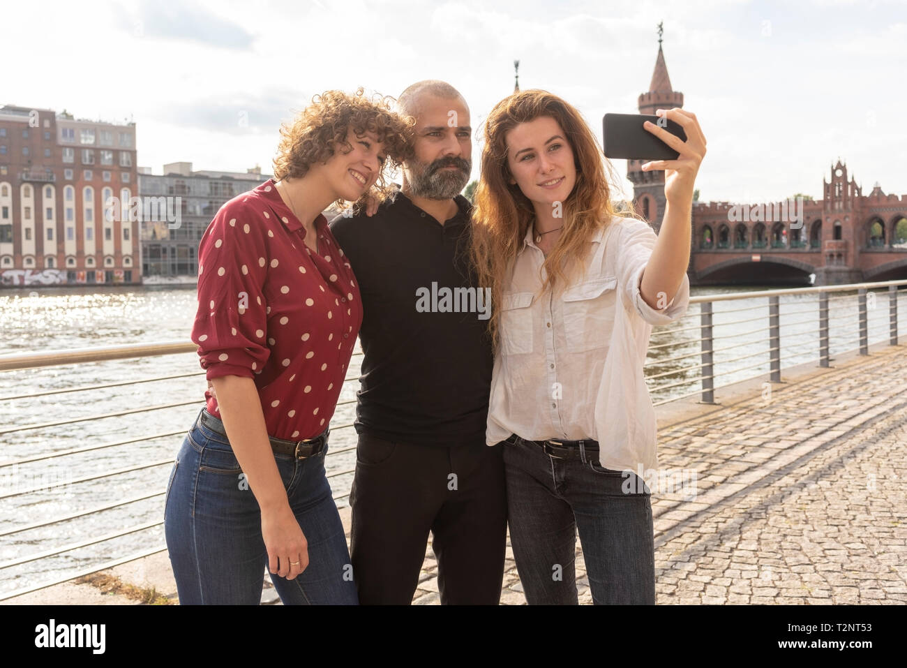 Uomo e donna amici prendendo selfie con lo smartphone sul ponte, sul fiume e gli edifici in background, Berlino, Germania Foto Stock
