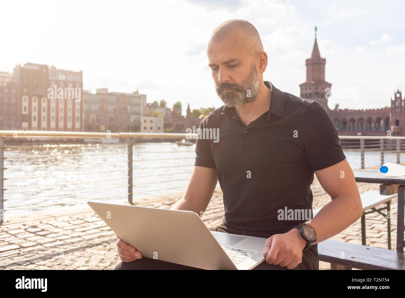Uomo che utilizza sul computer portatile sul ponte, sul fiume ponte Oberbaum ed edifici in background, Berlino, Germania Foto Stock