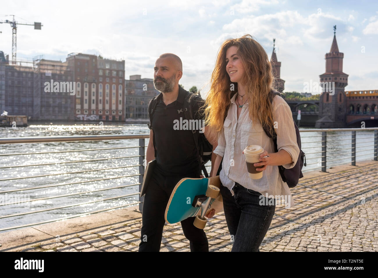 Uomo e donna amico con lo skateboard sul ponte, sul fiume ponte Oberbaum ed edifici in background, Berlino, Germania Foto Stock