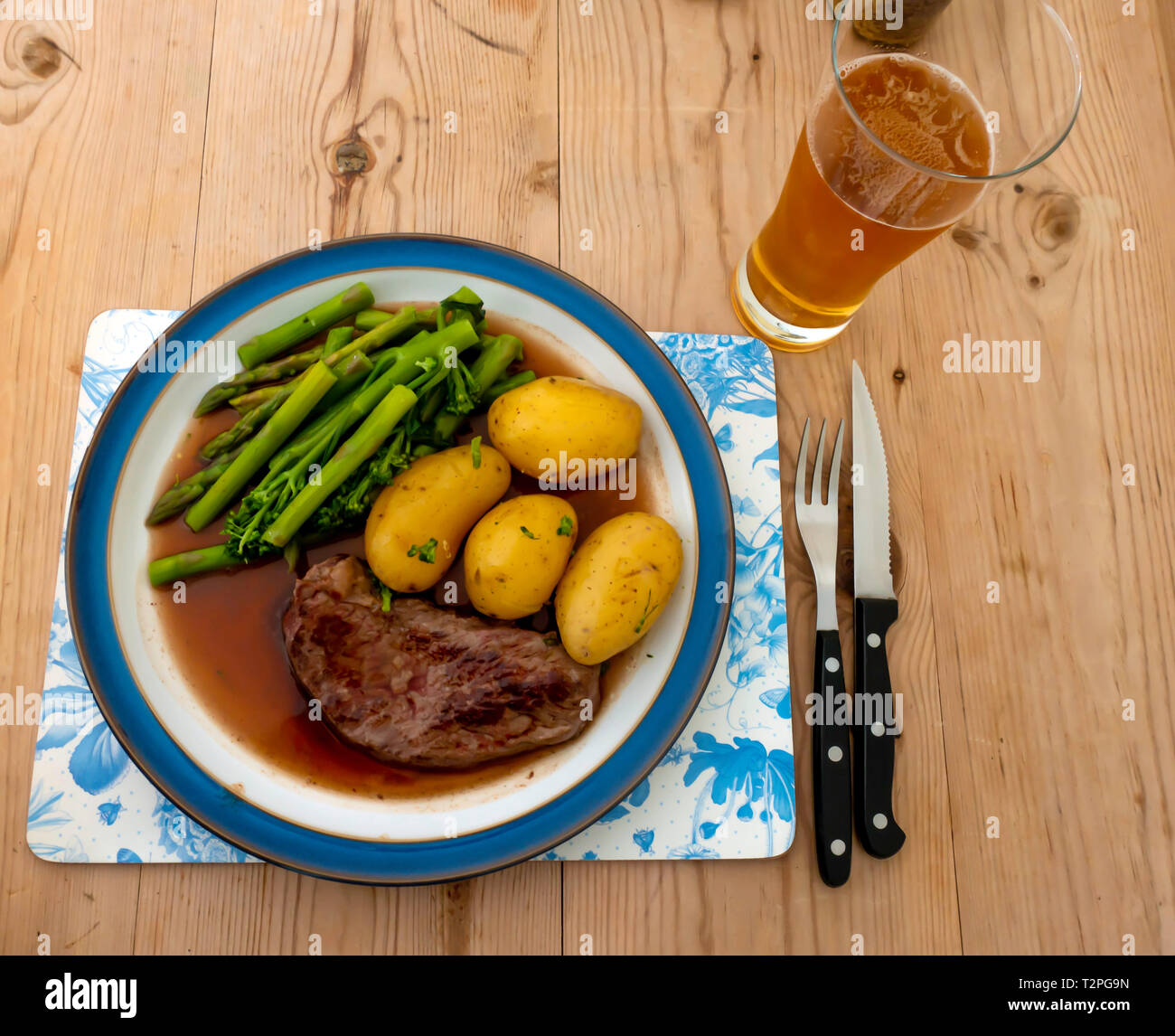 Un fritto Aberdeen Angus Scamone bistecca di manzo servita con patate di primizia, filetto di broccoli e asparagi e sugo di carne su un blu orlato piastra su una tavola di legno Foto Stock